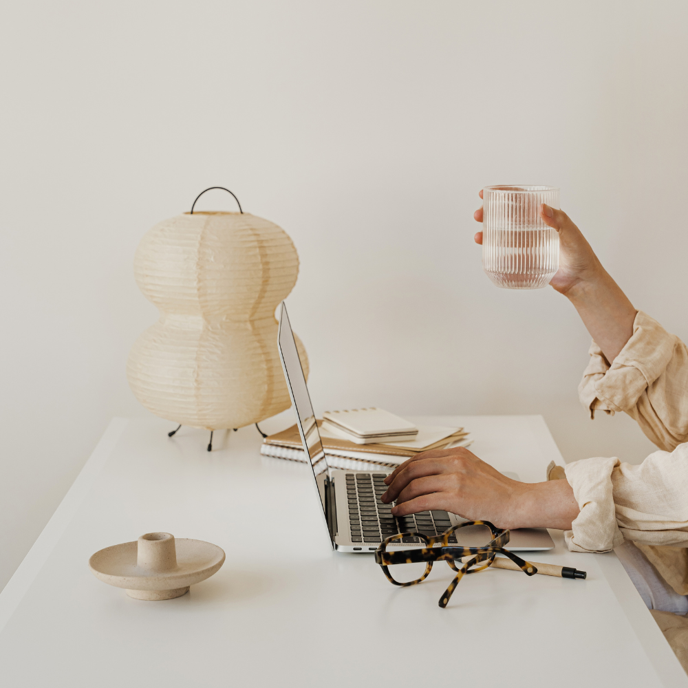 Person working on a laptop with a glass of water at a desk, surrounded by minimal decor.