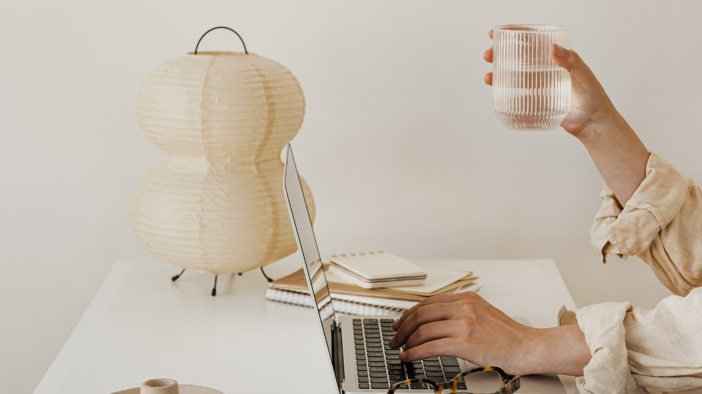 Person using a laptop with a cup in one hand and a lamp in the background