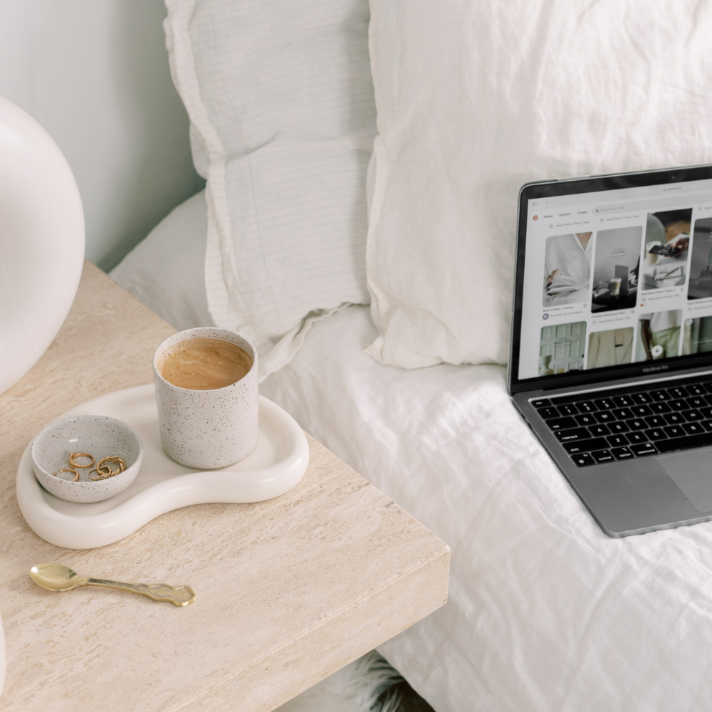 Laptop on a bed with a cup of coffee and small dish on a nightstand.