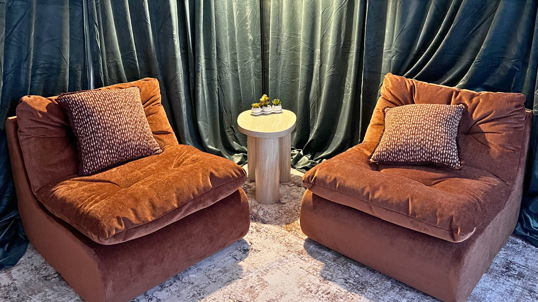 Two plush rust-brown velvet armchairs with patterned throw pillows, arranged symmetrically on a patterned rug in front of dark teal curtains, with a small round side table and a hanging lamp between them.